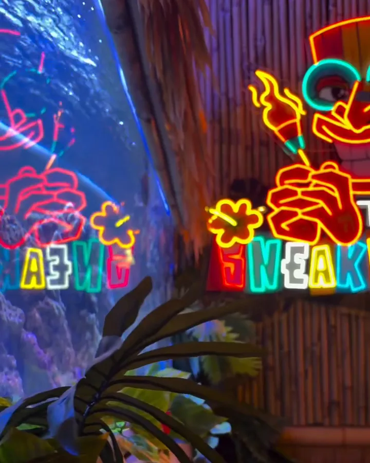 A corner of The Sneaky Tiki: the neon sign reflected in a reef tank, palms, and a bamboo rail